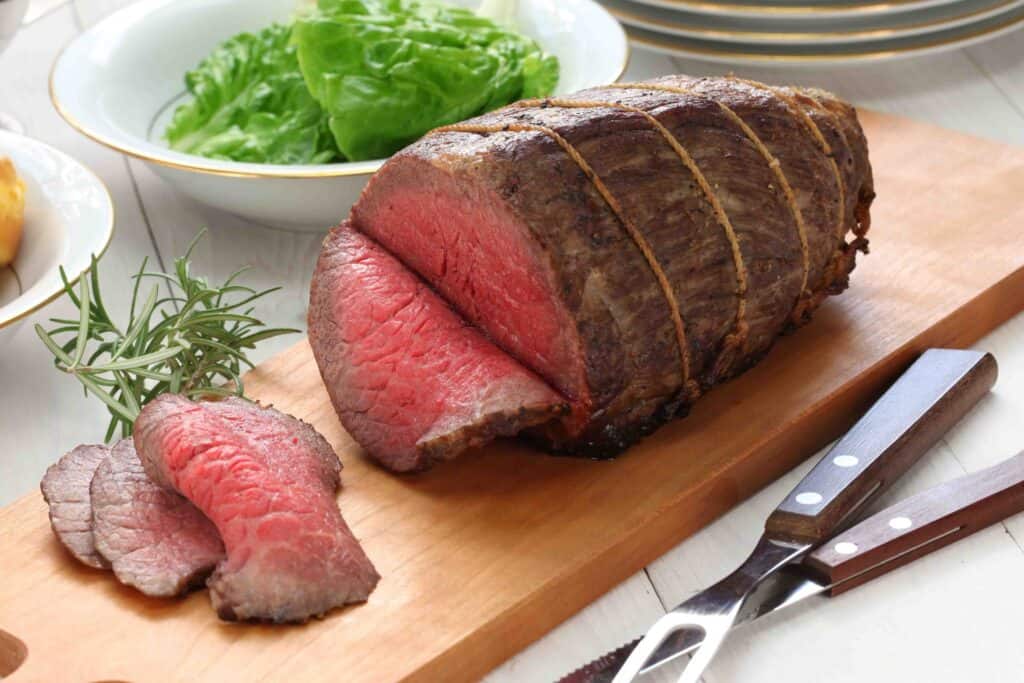 classic roast beef, tied and with three slices cut off the end, on a wooden board with a carving set in front and a bowl of lettuce behind it.